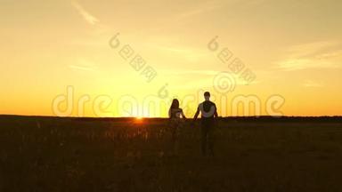 恋爱中的年轻夫妇牵着手穿过田野。 女孩和男人去看日落。 幸福的家庭散步